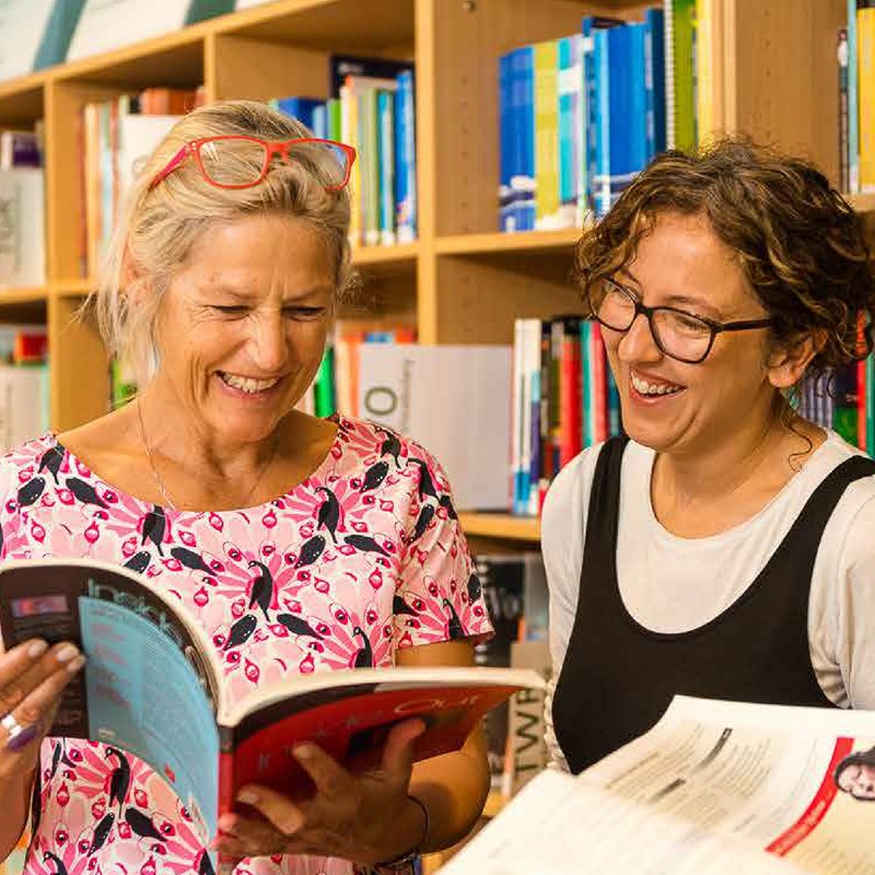 Two female teaching staff looking at books in a library laughing